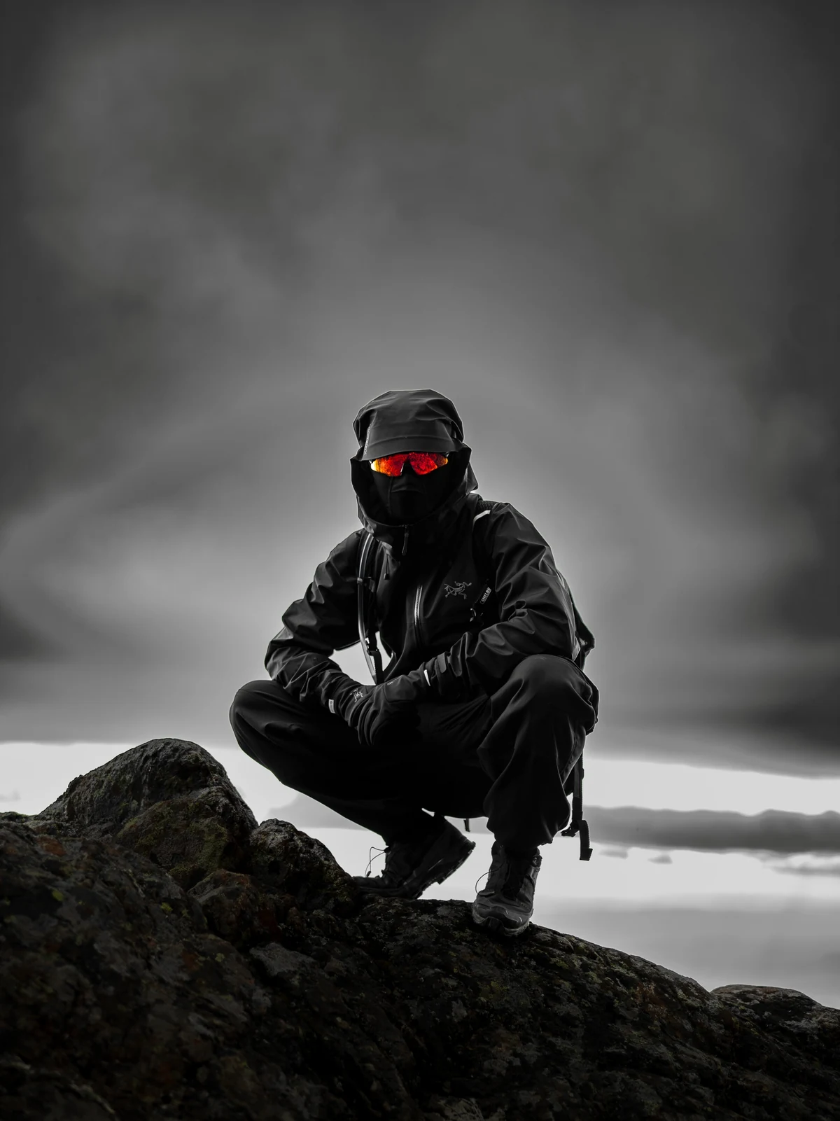 Person in black outdoor clothing and red mirrored sunglasses crouching on a rocky ridge beneath a cloudy sky. Shared by @ashridess, photographed by @mac.infocus.