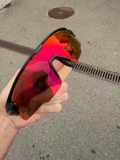 GORPED Strider sunglasses held up outdoors, the Ember lens reflecting buildings and trees