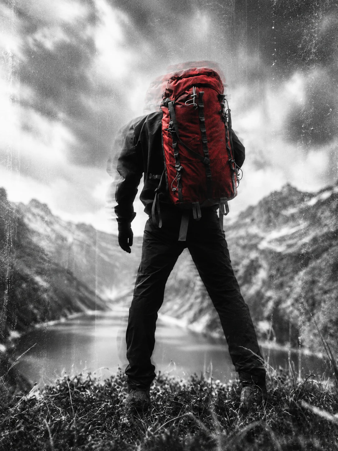 Hiker in black clothing with a red backpack, seen from behind against a grainy mountain landscape