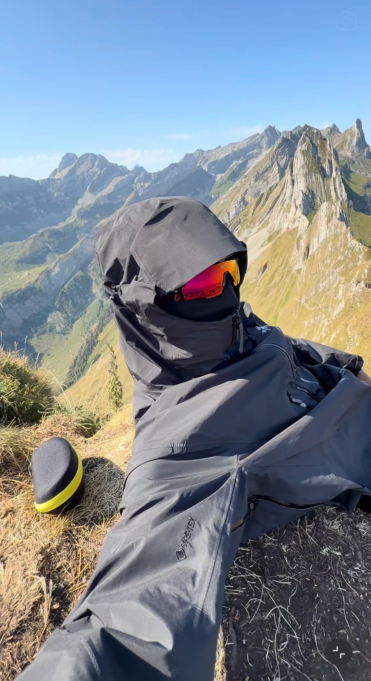 Hiker wearing a black hood and red mirrored Strider sunglasses on a grassy slope below alpine peaks, shared by @hikingcore1.
